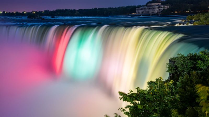 The Horseshoe Falls lit in pink, green and gold after dark, seen from the Canadian bank with the crest running the width of the frame