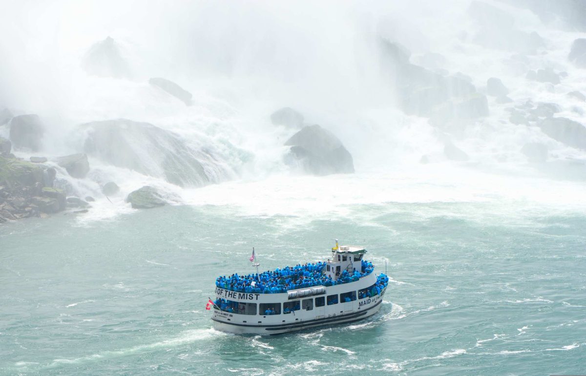 A Maid of the Mist boat crowded with blue ponchos below the American Falls, the mist wall rising behind it