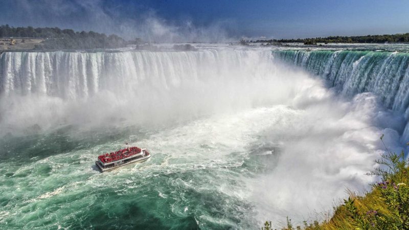 A Niagara City Cruises boat crowded with red ponchos in the Horseshoe Falls basin, dwarfed by the curtain of water