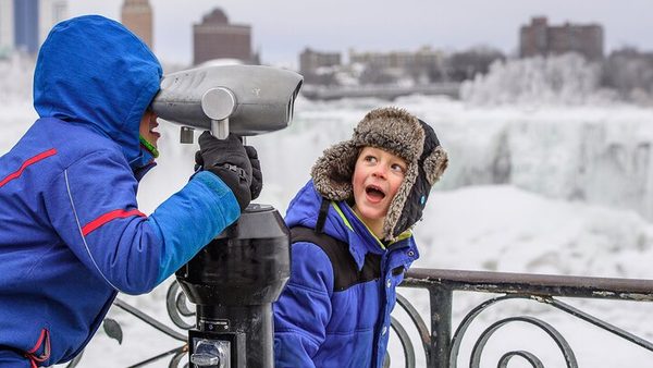 Two children in winter coats at a coin viewer on the Niagara Parkway with the frozen falls behind them