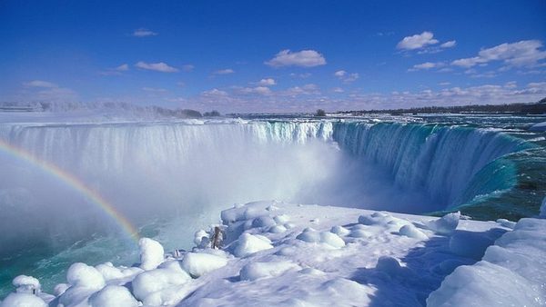 The Horseshoe Falls in winter from the Canadian bank, ice mounds at the base and a rainbow in the spray