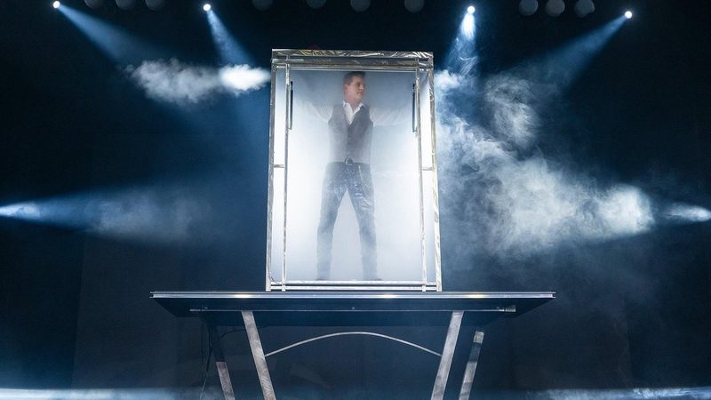 A performer standing inside a smoke filled glass cabinet raised on a steel table, lit by stage beams