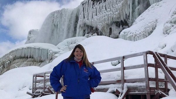 A visitor in a blue jacket at a snow covered railing below a wall of ice beside the frozen falls