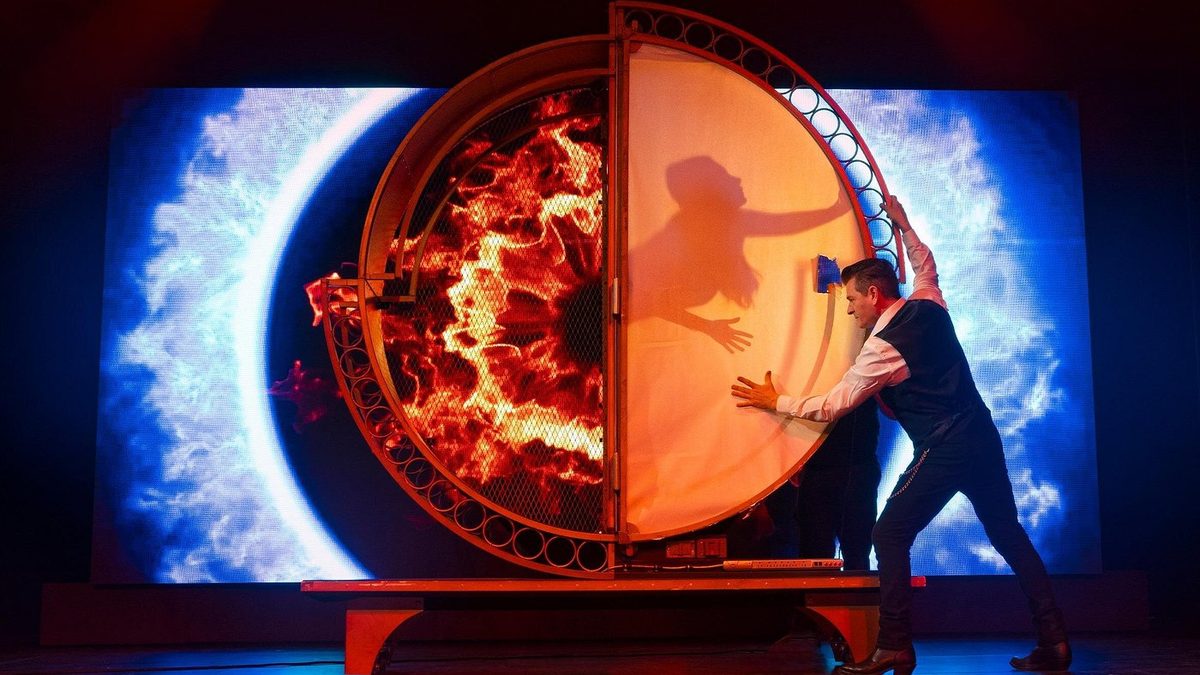A magician pressing shut a blazing circular illusion cabinet on the Niagara Magic Live stage, a silhouette visible inside it