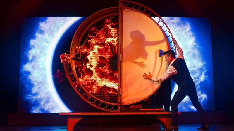 A magician pressing shut a blazing circular illusion cabinet on the Canadiana The Musical stage