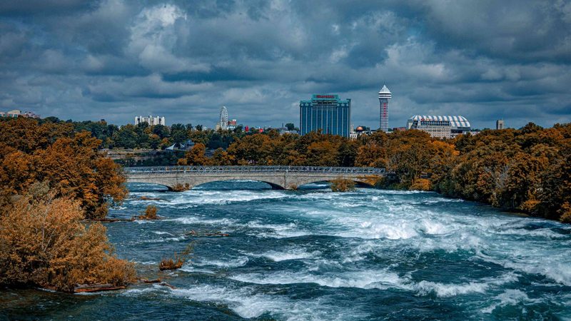 The upper Niagara River running fast past turning autumn trees, with the Niagara Falls, Ontario skyline and the Skylon Tower under heavy cloud behind
