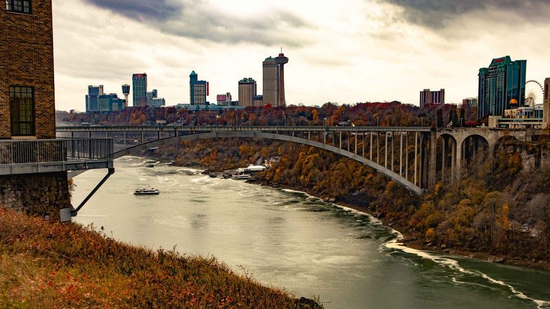 The Rainbow Bridge over the Niagara Gorge in autumn, with the Niagara Falls Ontario skyline beyond and a tour boat below