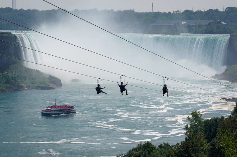 Three zipline riders flying toward the Horseshoe Falls, a red poncho boat on the river below