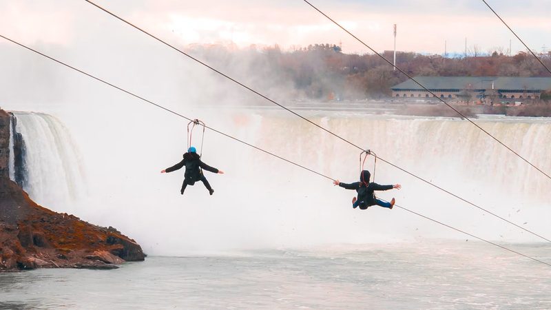 Two zipline riders with arms outstretched above the river, the Horseshoe Falls in autumn behind