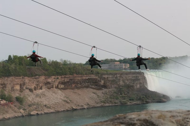 Three zipline riders in teal helmets above the gorge wall, the crest of the Horseshoe Falls behind